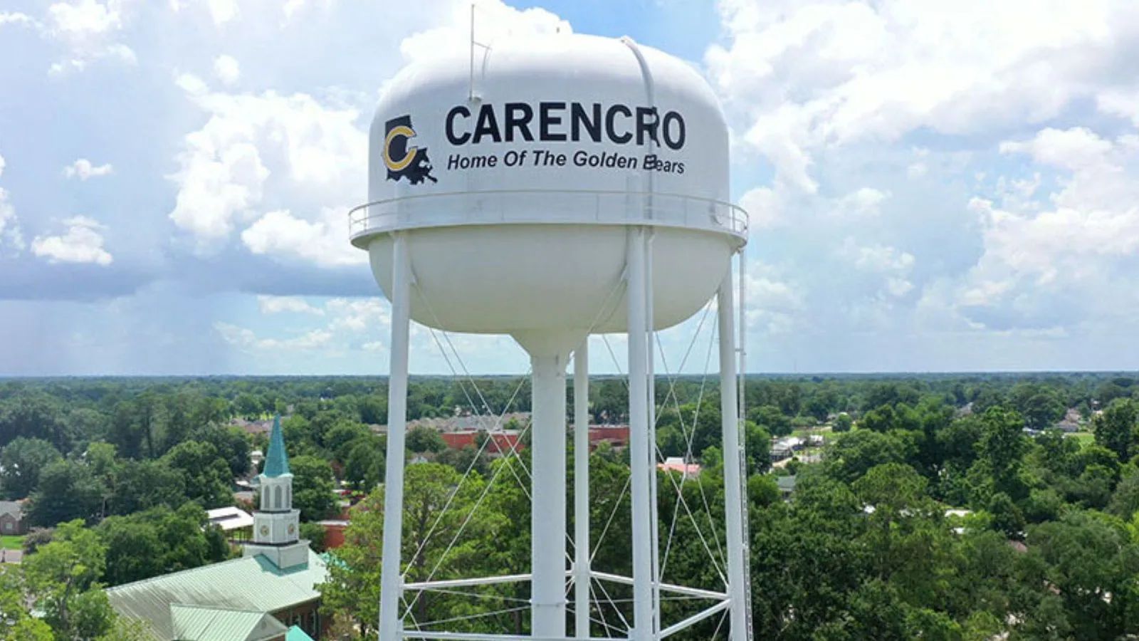 The Carencro water tower, Home of the Golden Bears, seen from the air above the treetops with a church spire below